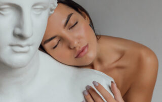A woman with a well-groomed face and body stands near the bust of a sculpture of a woman